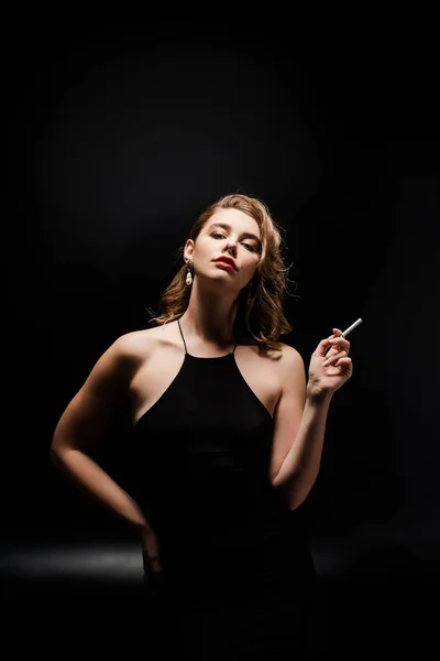 Attrayant femme en robe élégante tenant cigarette tout en posant sur fond noir — Photo de stock