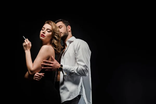 Bonito homem no branco camisa abraçando elegante, sexy mulher segurando cigarro isolado no preto — Fotografia de Stock