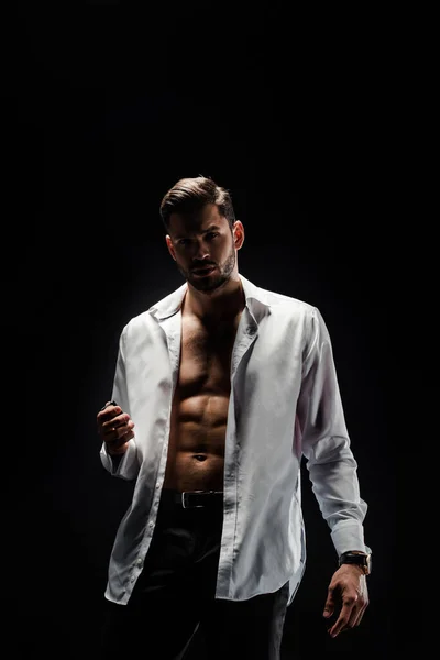 Сексуальный, мускулистый мужчина в белой расстегнутой рубашке, смотрящий на камеру, изолированную на черном — стоковое фото