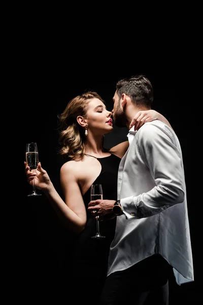 Bel homme en chemise déboutonnée et élégante, femme sexy tenant des verres de champagne et embrassant isolé sur noir — Photo de stock