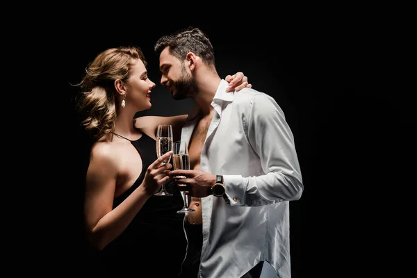 Sexy homem em branco desabotoado camisa e elegante, sedutor mulher segurando taças de champanhe isolado no preto — Fotografia de Stock