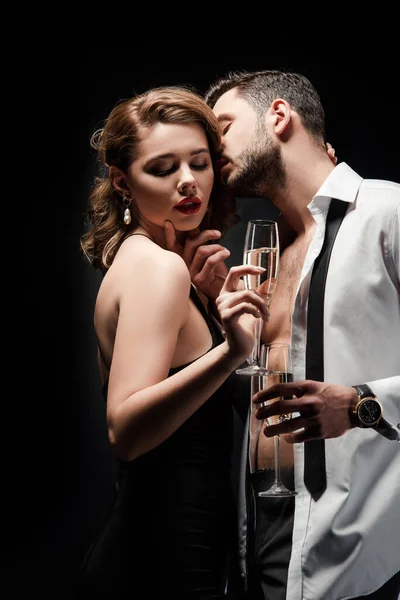 Sexy man in unbuttoned shirt kissing elegant, passionate girl holding champagne glass isolated on black — Stock Photo