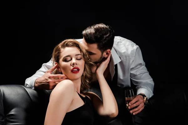 Handsome man holding champagne glass and kissing seductive woman sitting on sofa isolated on black — Stock Photo