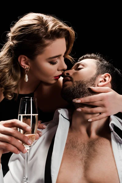 Attractive, elegant woman holding champagne glass and kissing handsome man isolated on black — Stock Photo