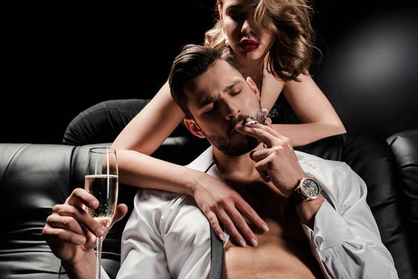 Donna sexy toccare bello, uomo sicuro seduto sul divano con bicchiere di champagne e fumare su sfondo nero — Foto stock