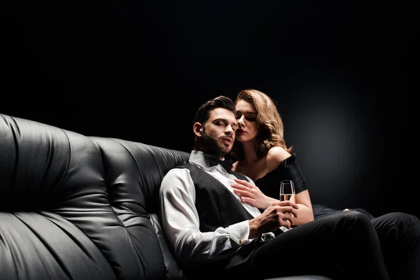 Attractive, sexy woman hugging handsome, elegant man sitting on leather sofa with champagne glass on black — Stock Photo