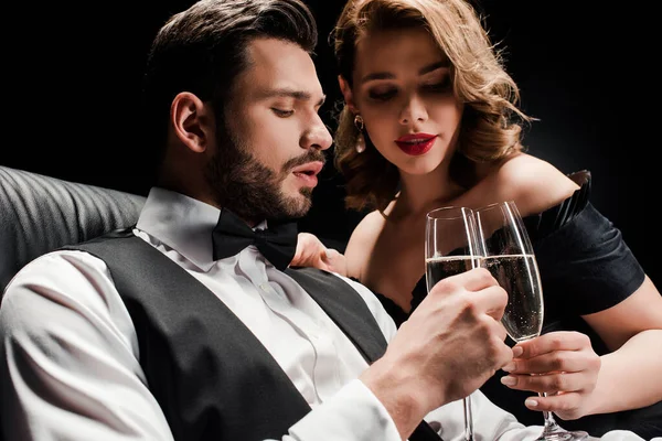 Atractiva mujer y elegante hombre tintineo copas de champán aislado en negro - foto de stock