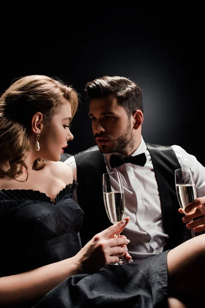 Sexy mujer y elegante hombre sosteniendo copas de champán y mirándose el uno al otro sobre fondo negro - foto de stock