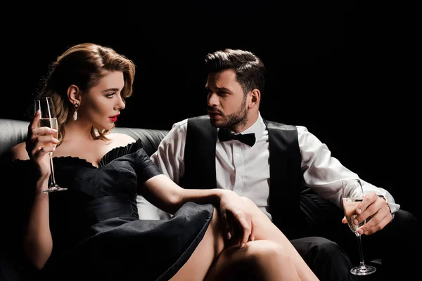 Elegante hombre y mujer sentados en un sofá de cuero con copas de champán y mirándose aislados en negro - foto de stock