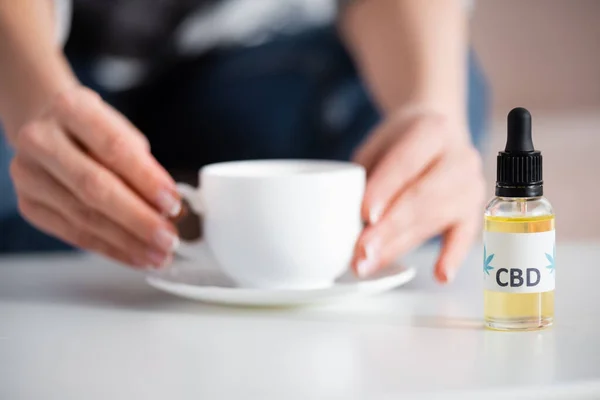 Vista cortada de mulher madura tocando xícara de chá perto de garrafa com letras cbd — Fotografia de Stock