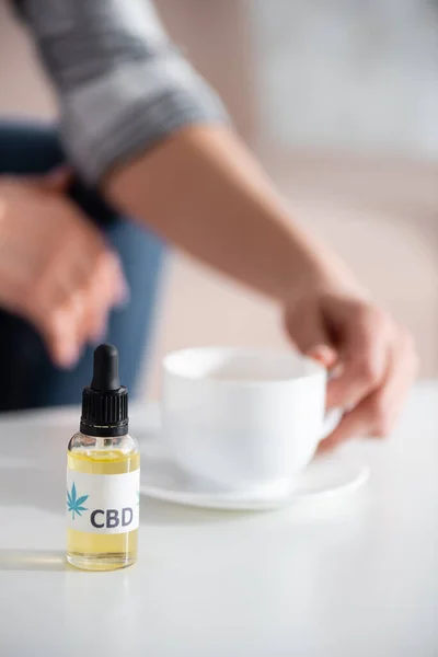 Selective focus of bottle with cbd lettering near mature woman touching cup of tea — Stock Photo