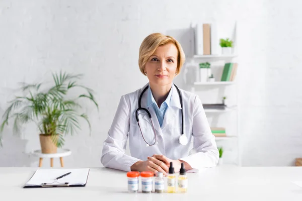 Mature doctor sitting at desk with bottles of cbd near clipboard — Stock Photo