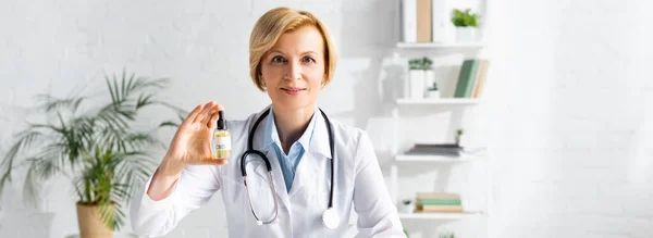 Panoramic shot of mature doctor in white coat holding bottle with cbd lettering in clinic — Stock Photo