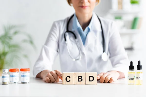 Vista cortada do médico em casaco branco tocando cubos de madeira com letras cbd perto de drogas — Fotografia de Stock