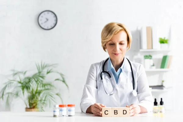 Doctor in white coat touching wooden cubes with cbd lettering near bottles with drugs — Stock Photo