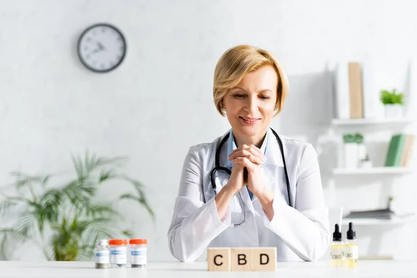 Happy doctor in white coat looking at cubes with cbd lettering near bottles with drugs — Stock Photo