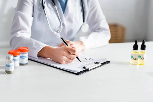 Vista cortada do médico que escreve a prescrição perto de garrafas com cannabis medicinal e cbd — Fotografia de Stock