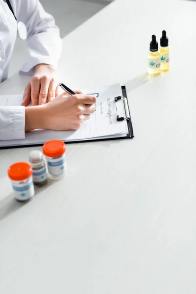 Vista recortada de médico que escribe prescripción cerca de botellas con cannabis medicinal y cbd en la mesa - foto de stock