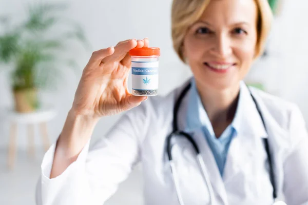 Selective focus of cheerful and mature doctor in white coat holding bottle with medical cannabis lettering — Stock Photo