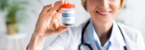 Prise de vue panoramique du médecin gai et mature en manteau blanc tenant bouteille avec lettrage de cannabis médical — Photo de stock