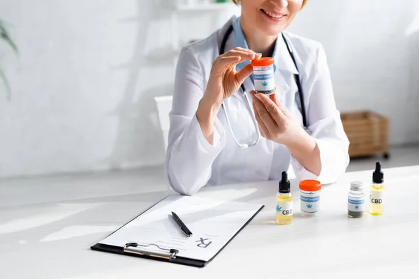 Cropped view of happy and mature doctor in white coat holding bottle with medical cannabis lettering — Stock Photo