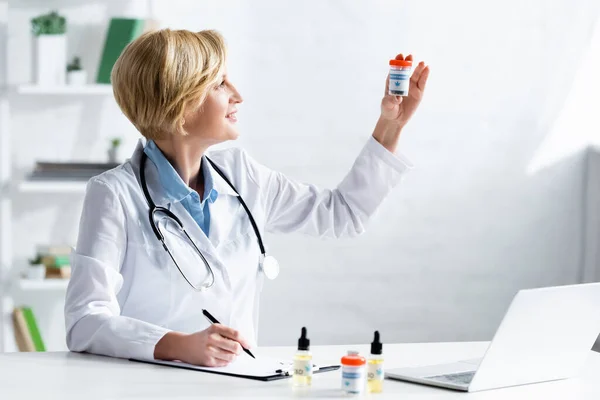 Médecin heureux et mature en manteau blanc regardant la bouteille avec lettrage de cannabis médical — Photo de stock