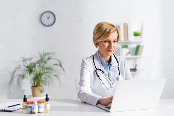 Mature doctor using laptop near bottles with medical cannabis and cbd lettering — Stock Photo