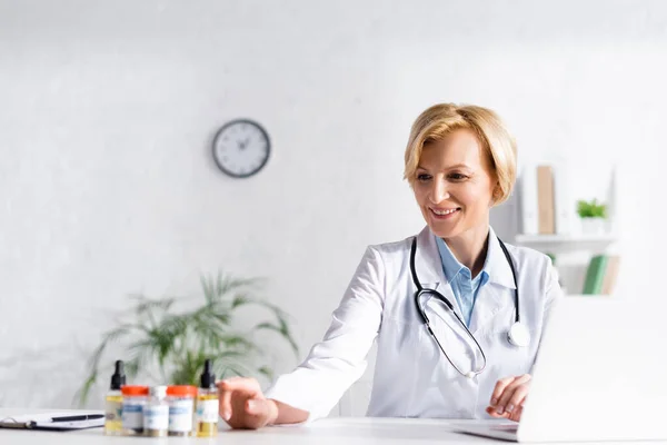 Médico feliz cerca de la computadora portátil y botellas con letras de cannabis medicinal - foto de stock