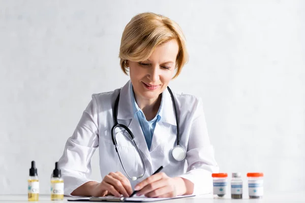 Selective focus of mature doctor writing prescription near bottles with medical cannabis and cbd lettering — Stock Photo