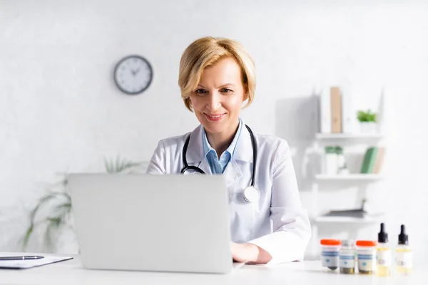 Glücklicher Arzt mit Laptop in der Nähe von Flaschen mit medizinischem Cannabis und cbd — Stockfoto