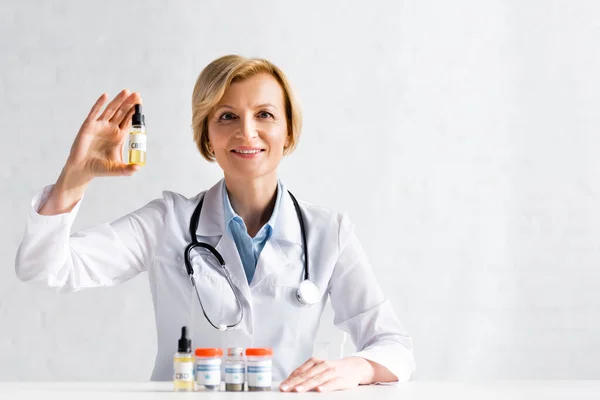 Cheerful and mature doctor in white coat holding bottle with cbd lettering in clinic — Stock Photo