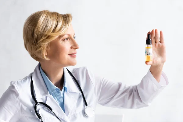 Médico feliz y maduro en blanco abrigo mirando botella con aceite y letras cbd en la clínica - foto de stock