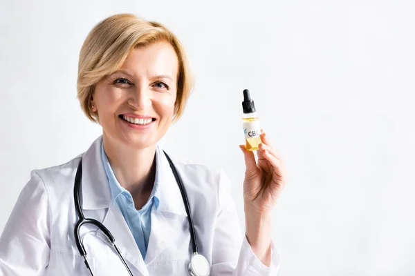 Happy and mature doctor in white coat holding bottle with oil and cbd lettering in clinic — Stock Photo