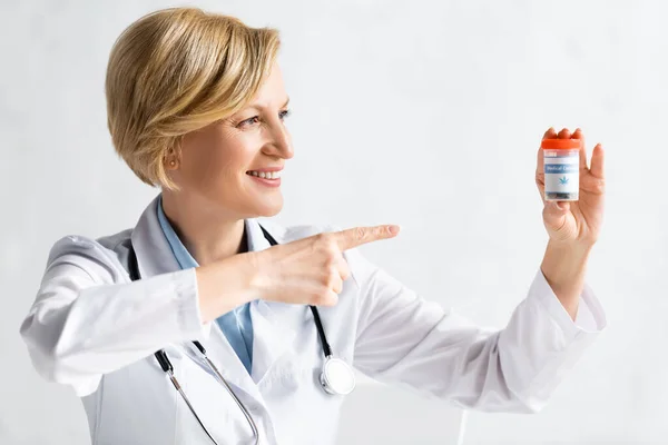 Happy and mature doctor in white coat pointing with finger at bottle with oil and cbd lettering in clinic — Stock Photo
