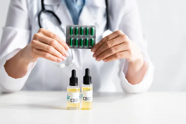 Обрезанный вид врача в белом халате, держащего пузырь пакет с таблетками возле бутылок с надписью CBD — стоковое фото