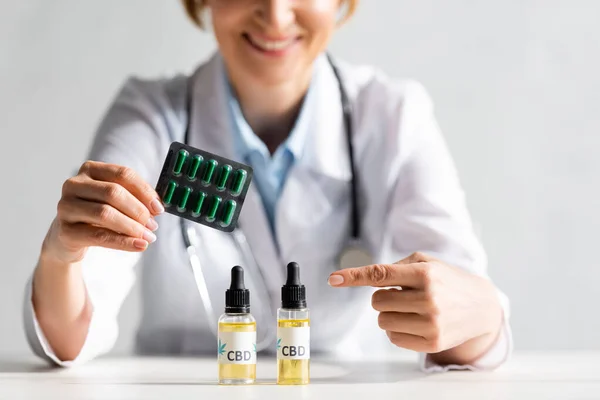 Cropped view of happy and mature doctor in white coat pointing with finger at bottle with cbd lettering and holding blister pack with pills in clinic — Stock Photo
