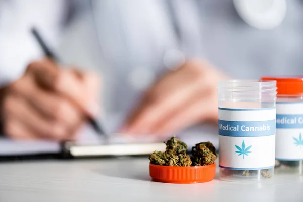Selective focus of dried weed and bottles with medical cannabis lettering near doctor writing prescription — Stock Photo