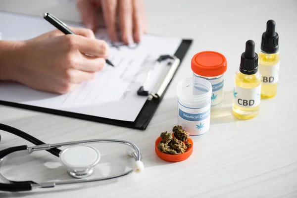 Ausgeschnittene Ansicht eines Arztes, der in der Nähe von getrocknetem Unkraut Rezepte schreibt, Flaschen mit cbd und medizinischem Cannabis-Schriftzug — Stockfoto