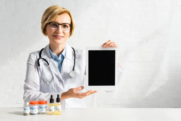 Mature doctor holding digital tablet with blank screen near bottles with cbd and medical cannabis lettering in clinic — Stock Photo