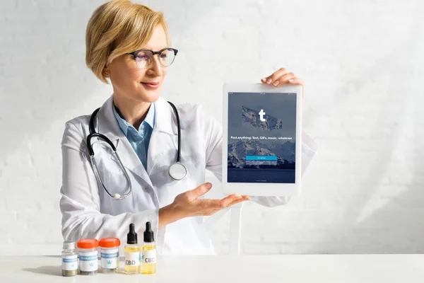 KYIV, UKRAINE - MARCH 5, 2020: mature doctor in white coat holding digital tablet with tumblr app near bottles with cbd and medical cannabis lettering — Stock Photo