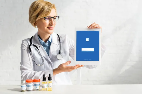 KYIV, UKRAINE - MARCH 5, 2020: mature doctor in white coat holding digital tablet with facebook app near bottles with cbd and medical cannabis lettering — Stock Photo