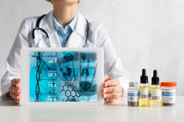Cropped view of mature doctor in white coat holding digital tablet with medical app near bottles with cbd and cannabis lettering — Stock Photo