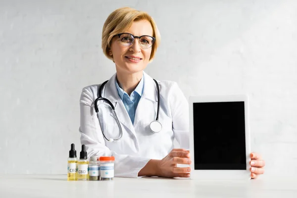 Mature doctor in white coat holding digital tablet with blank screen near bottles with cbd and medical cannabis lettering in clinic — Stock Photo