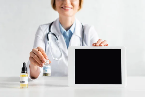 Vista recortada de médico feliz y maduro de capa blanca sosteniendo tableta digital con pantalla en blanco y botella con letras de cannabis medicinal - foto de stock