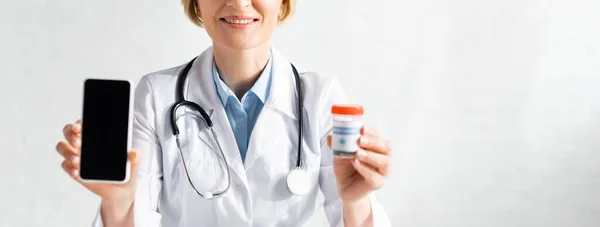 Panoramic shot of happy and mature doctor in white coat holding smartphone with blank screen and bottle with medical cannabis lettering — Stock Photo