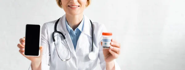 Prise de vue panoramique du médecin heureux et mature en manteau blanc tenant smartphone avec écran vierge et bouteille avec lettrage de cannabis médical — Photo de stock