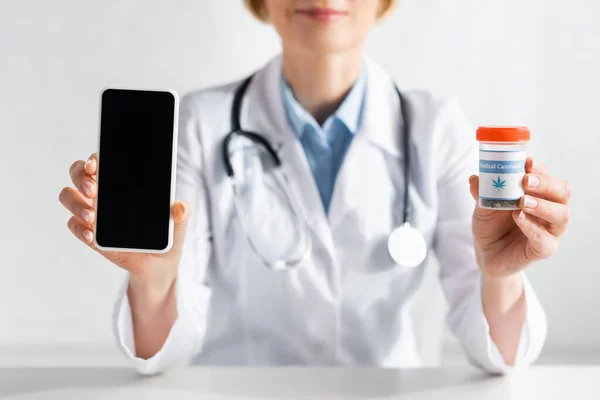 Cropped view of mature doctor in white coat holding smartphone with blank screen and bottle with medical cannabis lettering — Stock Photo