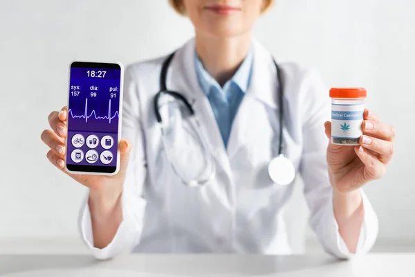 Cropped view of mature doctor in white coat holding smartphone with heartbeat rate and bottle with medical cannabis lettering — Stock Photo
