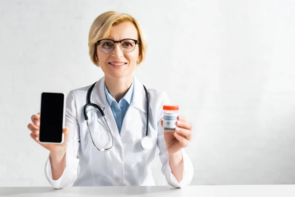 Doctor feliz y maduro en bata blanca sosteniendo teléfono inteligente con pantalla en blanco y botella con letras de cannabis medicinal - foto de stock