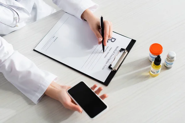 Cropped view of doctor writing prescription near bottles with cbd and medical cannabis while holding smartphone with blank screen — Stock Photo
